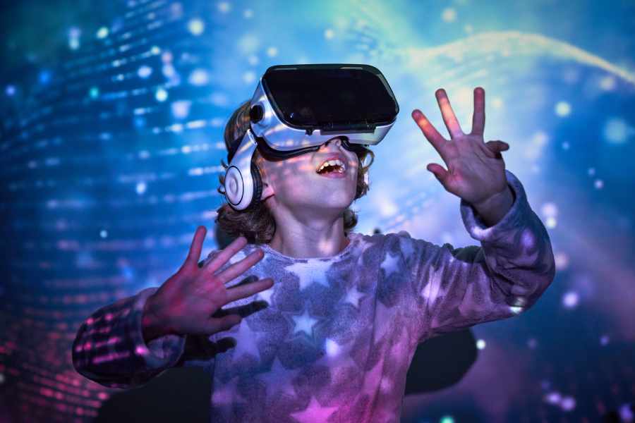 Joyful little child with blond hair in warm nightwear and VR goggles smiling and looking up with astonishment while exploring cyberspace in room with glowing neon lights