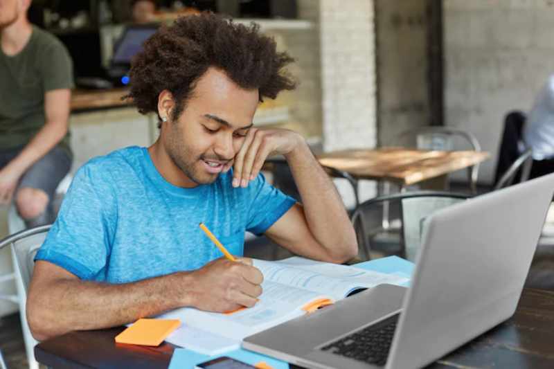 hardworking african american graduate student dressed casually making notes with pencil textbook while looking information course paper surfing high speed internet laptop pc 273609 7539