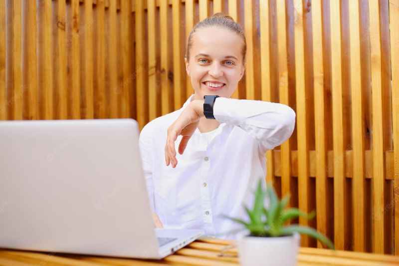 outdoor shot smiling woman wearing white shirt glasses sitting office with wooden walls working laptop using smart watch expressing positive emotions 176532 20903 1