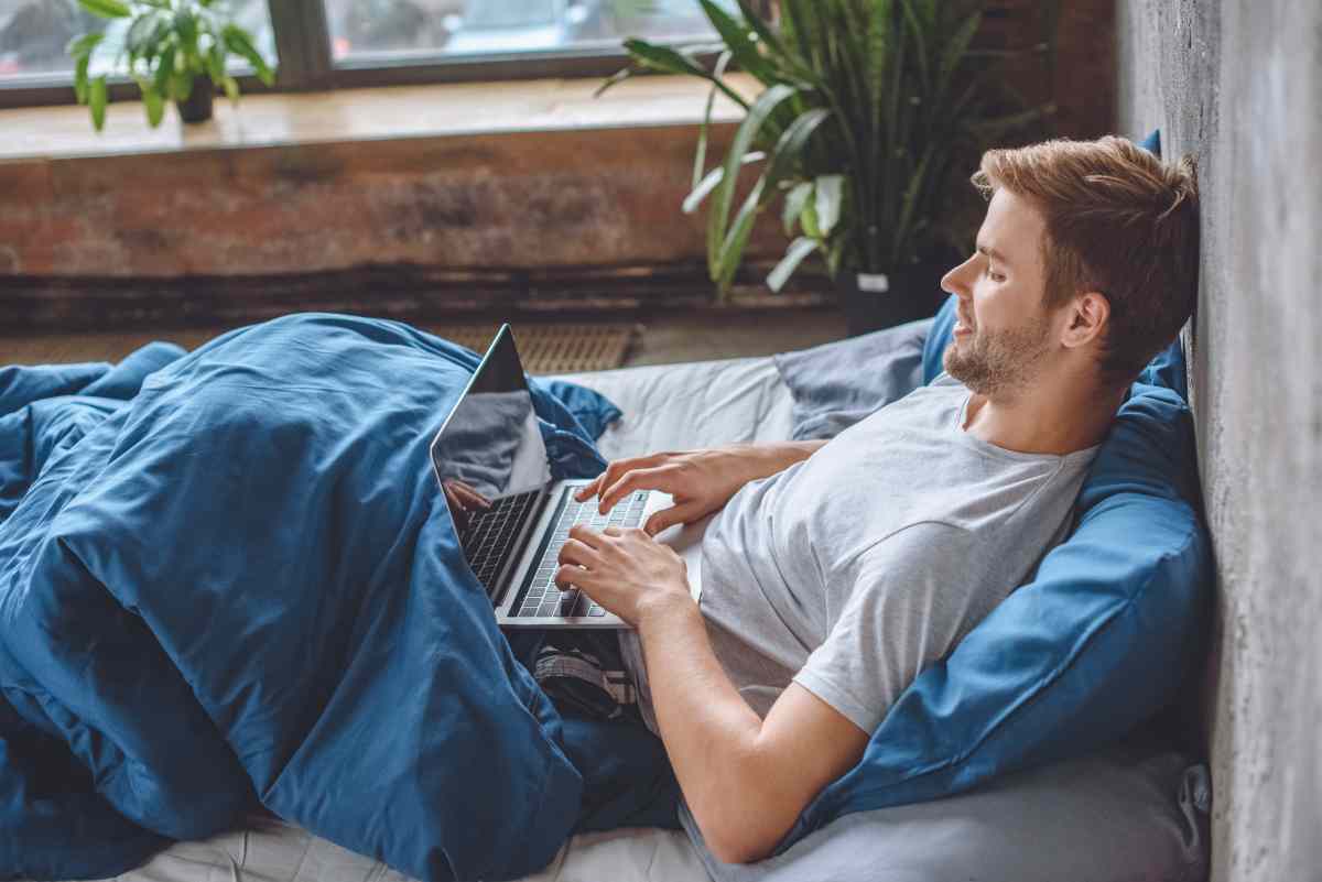 smiling man in bed using laptop with booking websi 2022 12 16 21 28 16 utc 1